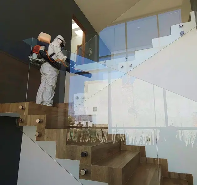 Fumigador profesional con traje blanco y equipo completo de fumigación en escaleras de madera con barandilla de cristal, interior de casa moderna con paredes blancas; Fumigaciones Potosí en San Luis Potosí